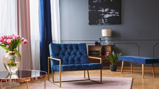 Salon avec un fauteuil bleu, un banc et un mur sombre. Les rideaux sont ouverts et un magnifique vase trône sur une table basse 