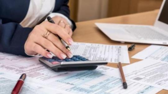 personne assise à son bureau devant son ordinateur, qui tient une calculatrice en main. Des feuilles sont éparpillées sur la table.